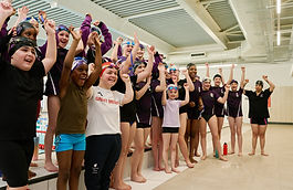 Maisie Summers-Newton and a group of diverse swimmers standing side by side indoors, raising their arms in celebration or cheer.