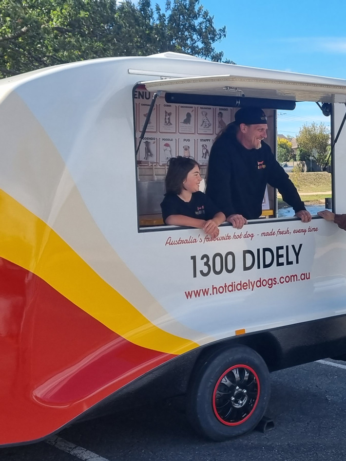 Small fibreglass food vending trailer, with side door up. Man and boy in hot didely dogs black uniforms standing inside food vending trailer with a customer standing outside