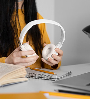 Hands holding white headphones resting on open books on a desk.