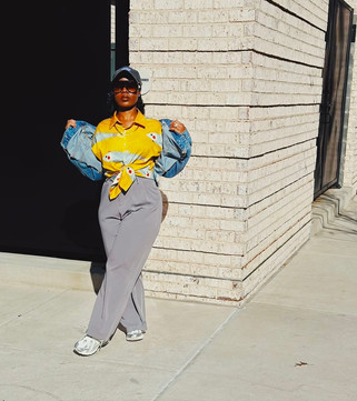 Mrs. VP rocking '90s streetwear with a yellow fish print shirt, grey trousers, and cropped denim jacket, posed against a white brick wall