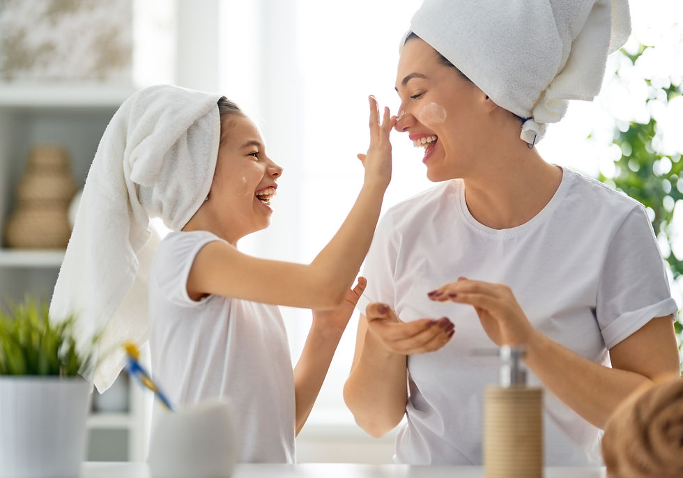 A mother and child both with hair up in white towels and wearing white shirts. Both are laughing as the child puts lotion on the mother's nose.