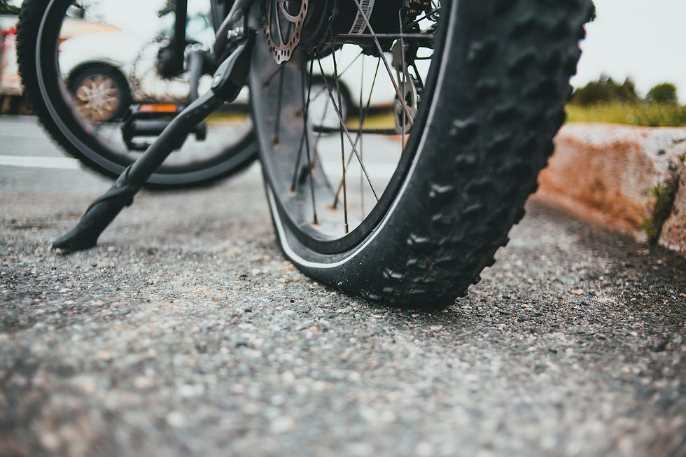 Close-up of a bicycle tire with a flat on a rough asphalt surface. Nearby, a blurred car tire is visible. The scene conveys a sense of inconvenience.