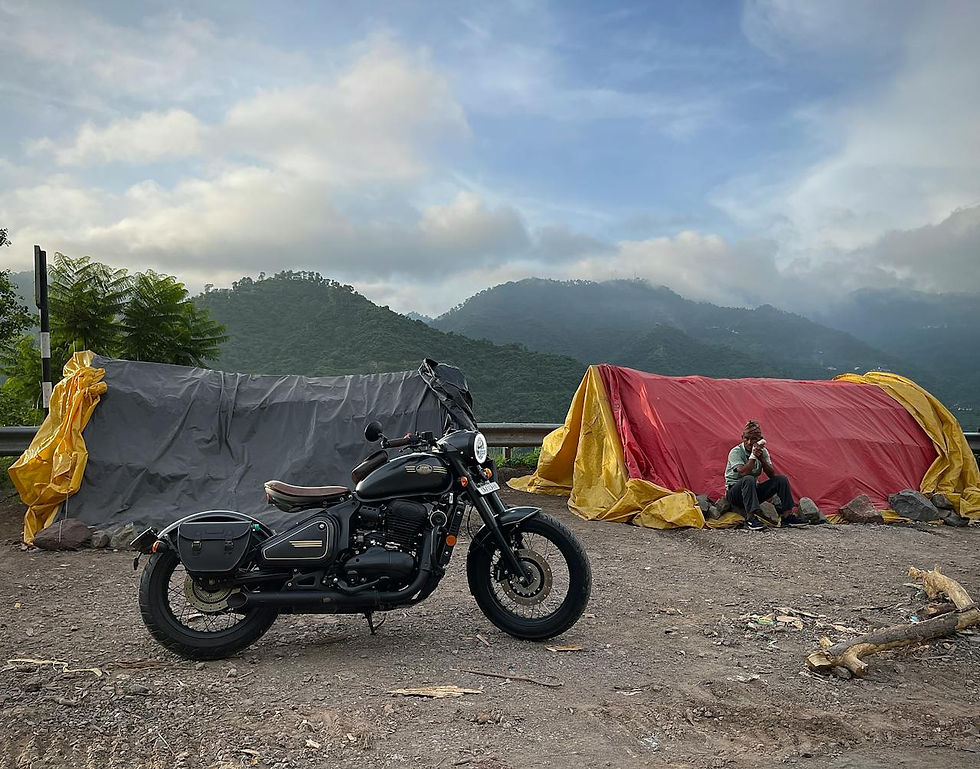 Motorcycle parked by a roadside tent with a man sitting beside. Misty hills in the background. Tents covered in yellow and red tarps.
