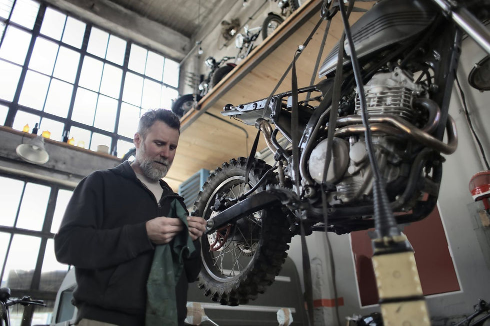 Mechanic inspects a motorcycle in a workshop with large windows. The room is industrial, and the man seems focused, holding a cloth.