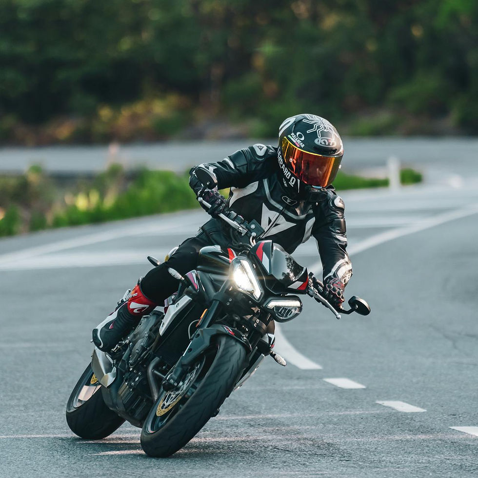 Motorcyclist in black gear with red visor leans into a curve on a road. The headlight is on. Background is blurred greenery.