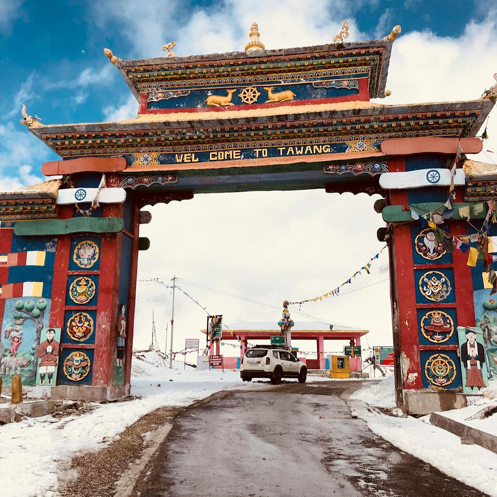 Colorful archway with Tibetan designs reads "WELCOME TO TAWANG." A car drives through snow. Overcast sky and vibrant prayer flags.