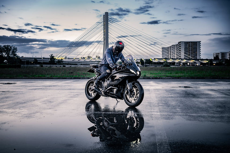 Motorcyclist in black gear on a parked bike, reflected in wet pavement. Modern bridge and buildings in the twilight background. Calm mood.