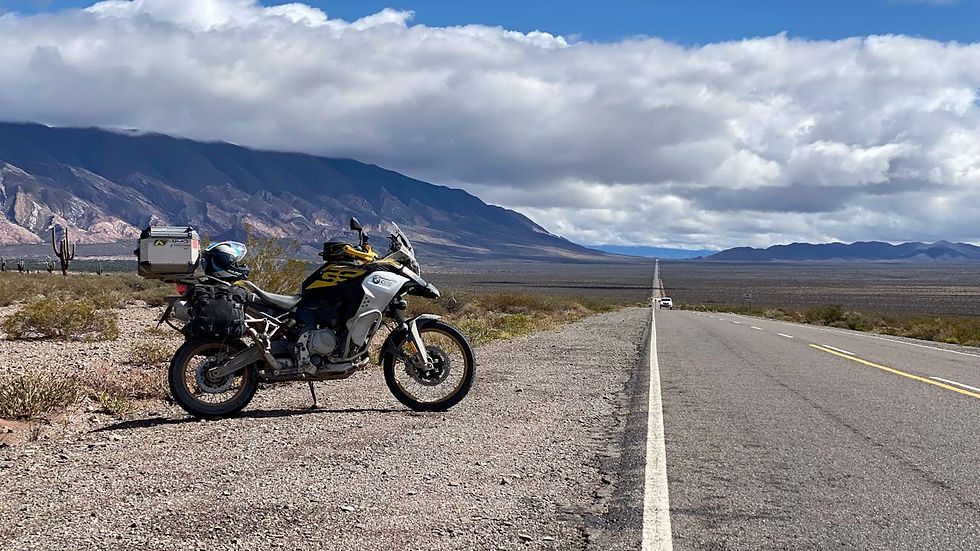Motorcycle parked beside a long road in a desert landscape with mountains under cloudy skies. A road sign and distant car visible.