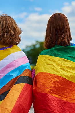 Two queer youth stand with their backs facing the camera, one is wearing the progress pride flag as a cape and the other is wearing a rainbow flag as a cape.