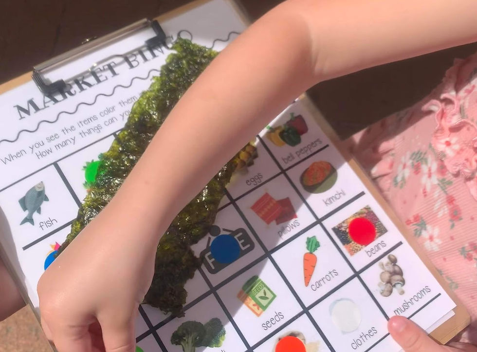 Child's arm pointing at a market bingo card on a clipboard, covered partly by green seaweed. The card has images and words like fish and seeds.
