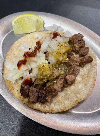 Taco with grilled meat, onions, and salsa on a metal plate. Lime wedge on the side. Background is dark.