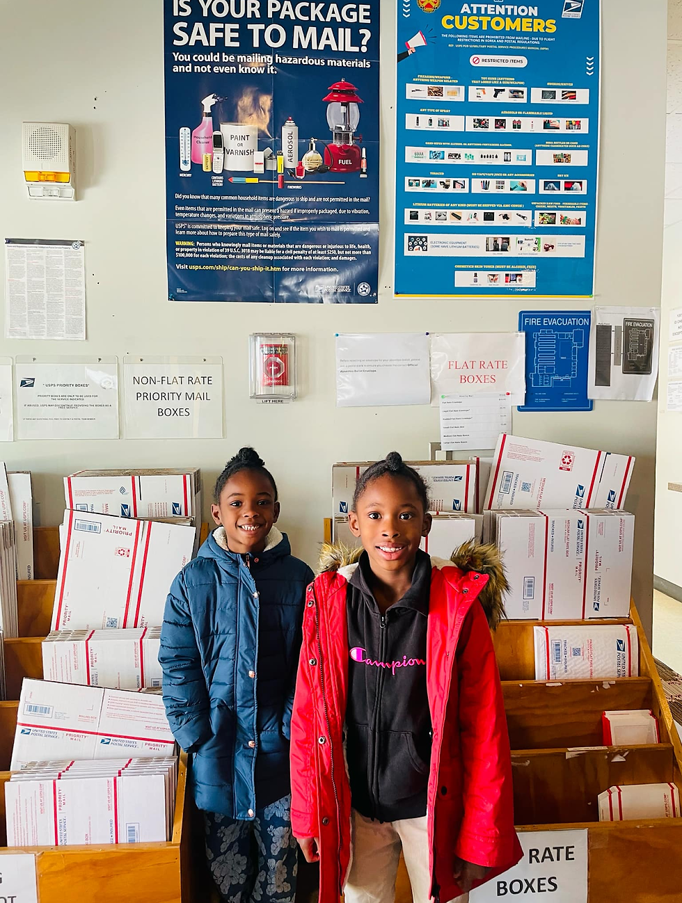 Two children smiling in a post office, surrounded by priority mail boxes. Posters about mailing safety and restrictions are on the wall.