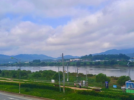 River scene with cloudy sky, distant mountains, and green landscape. A few people walk on a riverside path. Bridge and road in foreground.