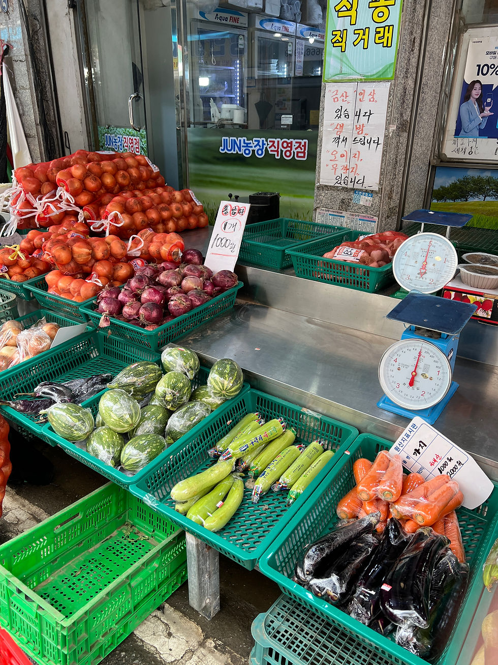 Vegetable market stall with green baskets of zucchini, eggplants, carrots, and melons. Signs with Korean text and scales visible.