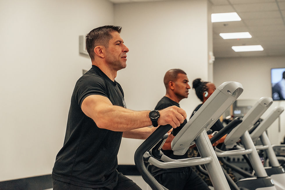Three people exercising on ellipticals in a gym, wearing black attire. The background features a plain wall and ceiling lights.