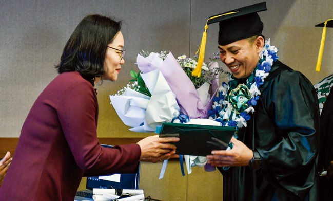 Man in graduation attire receives a flower bouquet and diploma from a woman in a red jacket. They are smiling in a celebratory setting.