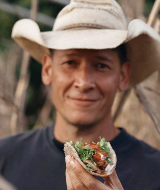 Man in a straw hat holding a loaded taco, smiling outdoors with greenery in the background. The setting is relaxed and rustic.