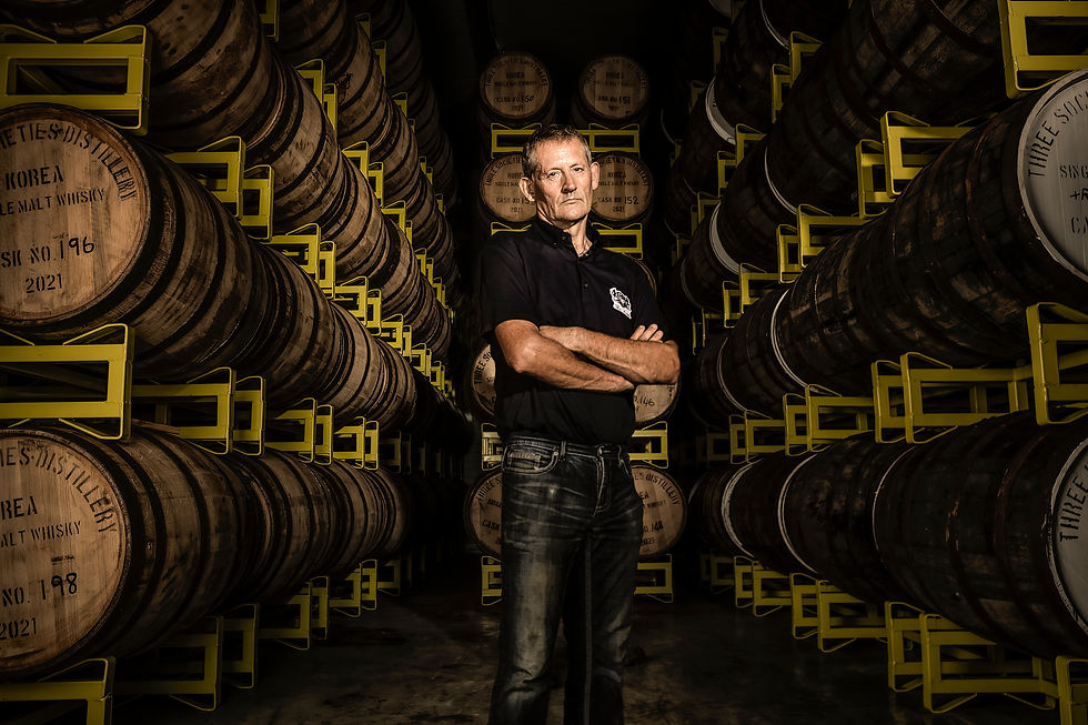 Man with arms folded across chest stands among barrels stamped with Three Societies Distillery in Aging room