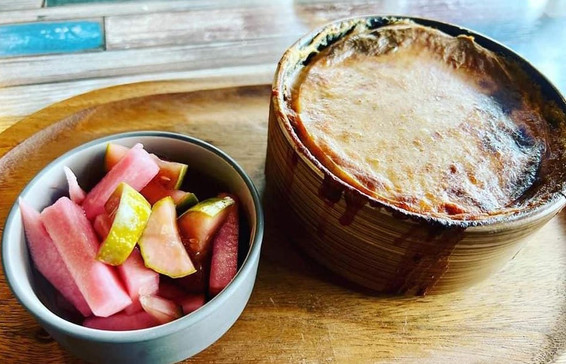 A pie in a wooden bowl with a golden crust sits beside a bowl of sliced pink pickles on a wooden tray. The setting has a casual vibe. Mousaka