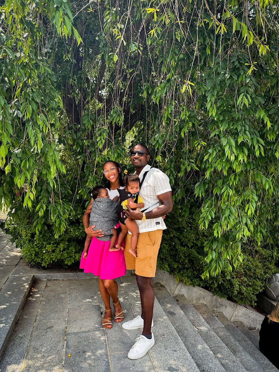 Smiling couple with two babies in carriers stand on stone steps, surrounded by lush green foliage. Woman wears a pink skirt.