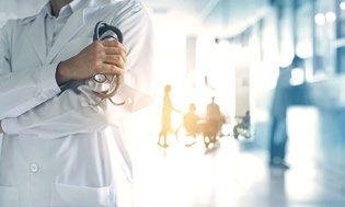 Doctor in a white coat holds a stethoscope, standing in a bright hospital corridor. Silhouettes of people and a wheelchair are visible in the background.