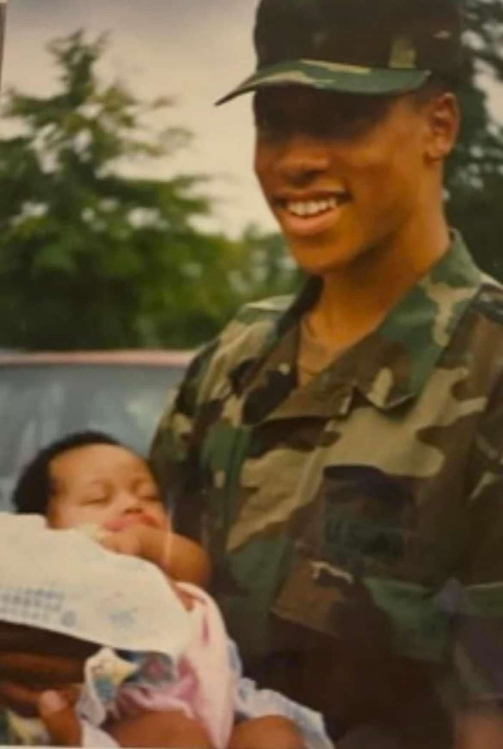 Smiling person in camouflage uniform holds a sleeping baby outdoors. Greenery in the background creates a serene mood.