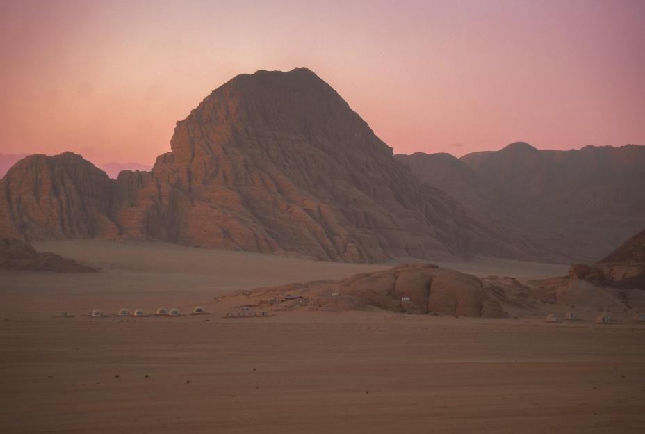 Desert landscape at sunset with a towering rock formation and small tents below. Sky is pink and purple, creating a serene and tranquil mood.