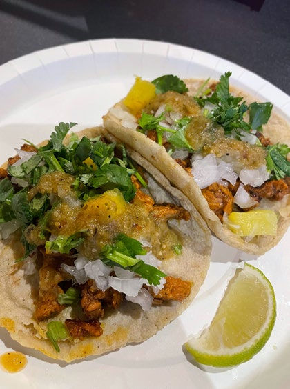 Two tacos with meat, cilantro, onions, and green salsa on a white plate. A lime wedge is beside the tacos on the plate.