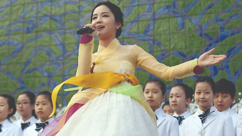 Young woman in colorful hanbok sings into a microphone, surrounded by children in white uniforms. Mosaic wall with green and blue in background.