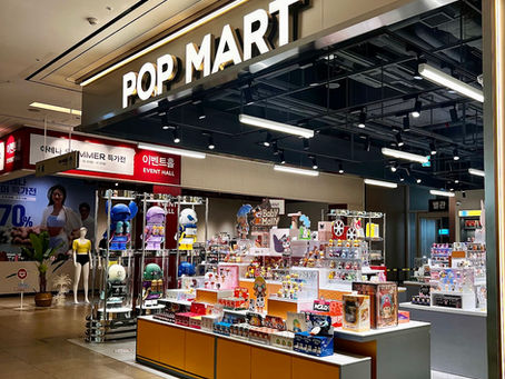 Pop Mart store interior with bright lights and toy displays. Prominent "POP MART" sign above. Colorful figurines on shelves create a playful mood.