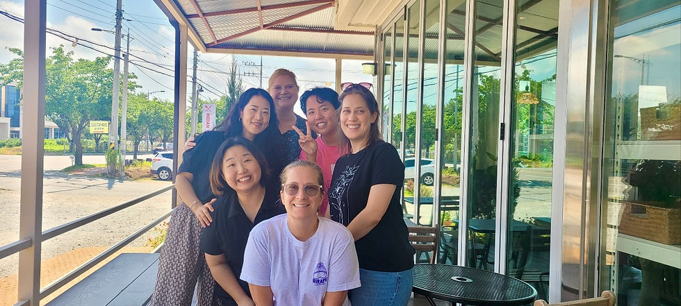 Smiling group of six people posing outside a café with glass walls. Sunny day, trees in background, casual attire, cheerful mood.