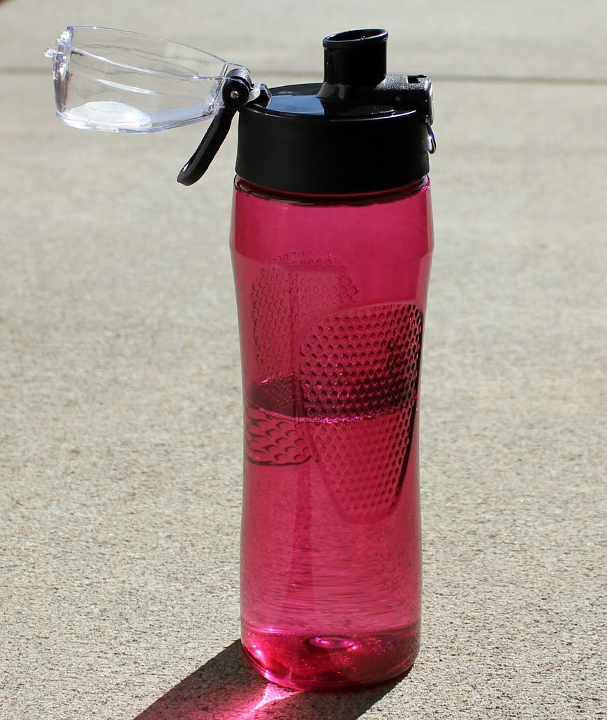 A pink water bottle with a black flip cap stands upright on concrete, partially filled and casting a shadow. Sunlit, outdoors.