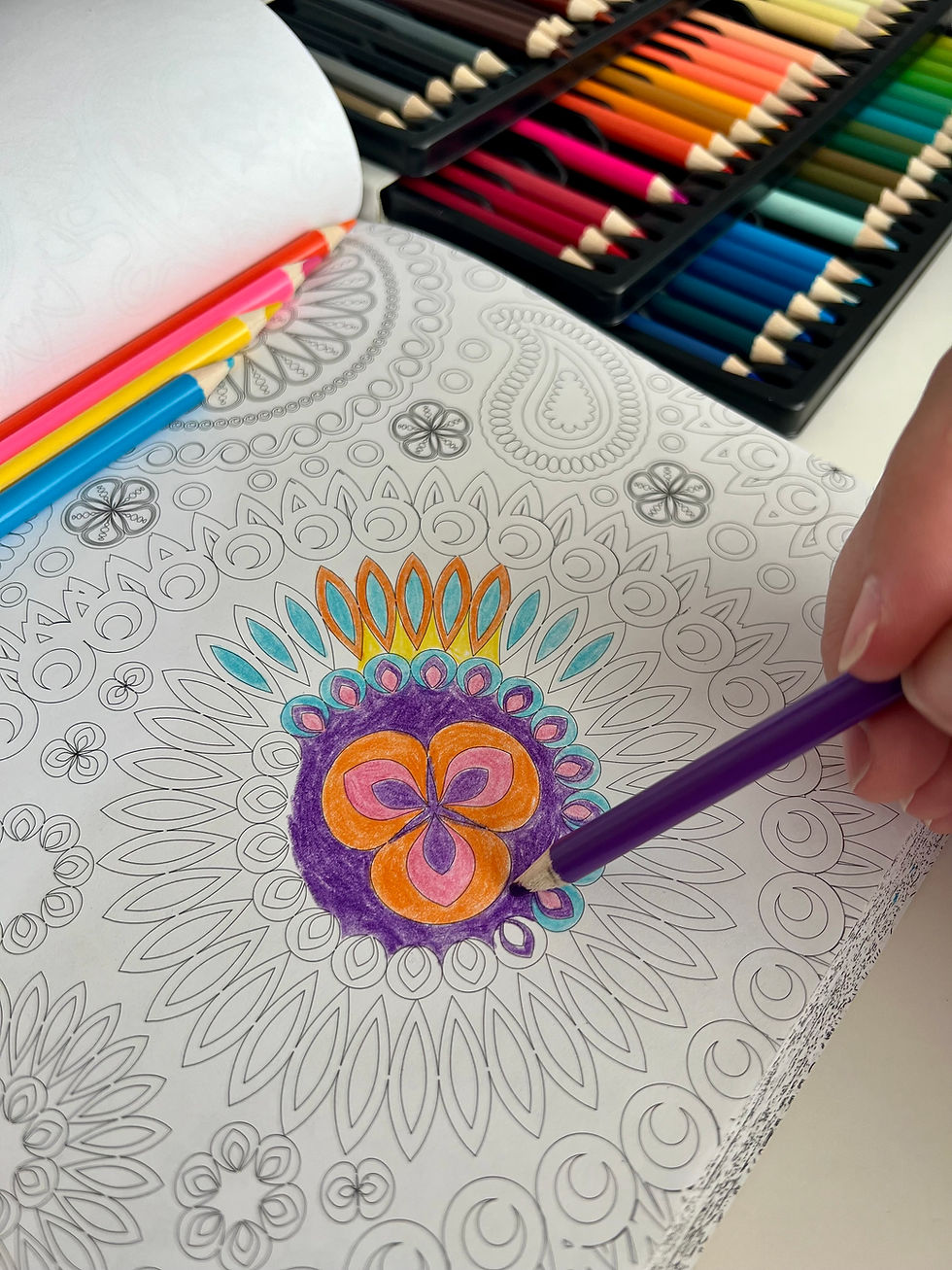 Hand coloring a detailed mandala with a purple pencil. Colorful pencils in a box nearby. Bright and intricate design fills the page.