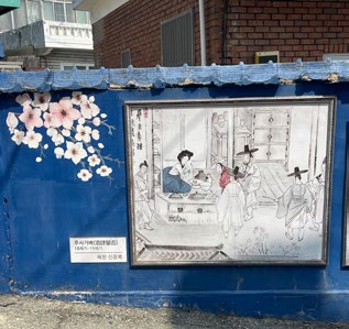 Framed traditional artwork on a blue wall with cherry blossoms. Depicts people in historical attire indoors. Text visible near the frame.