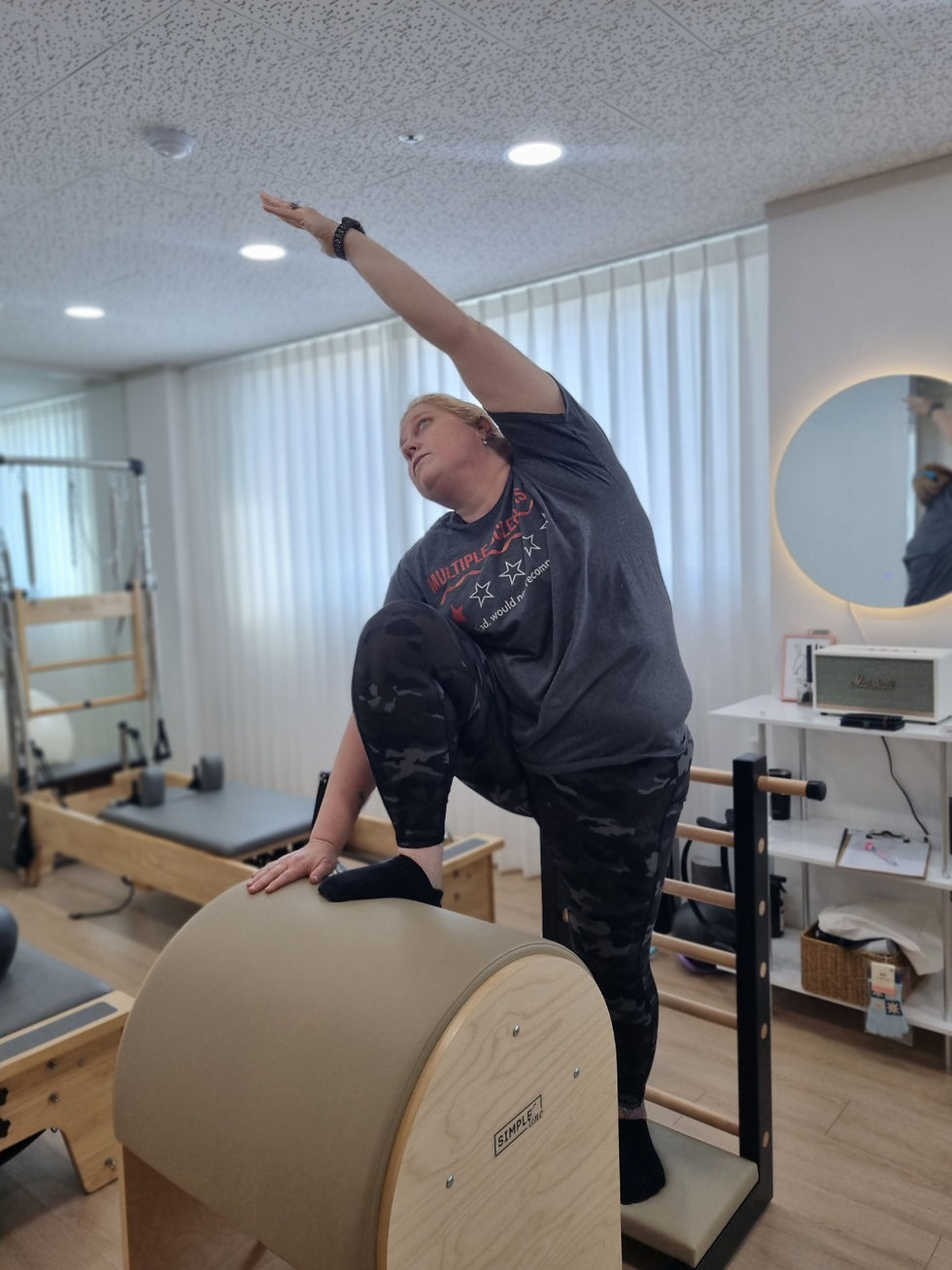 Woman in a gray shirt doing a stretch on Pilates equipment in a bright room with mirrors and white curtains, exuding focus and calm.