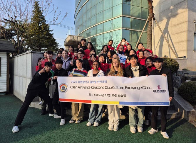 A group of people holding a banner outside a modern building. They're smiling, wearing casual clothes. Banner reads: "Osan Air Force Keystone Club Culture Exchange Class" with date December 10, 2024.