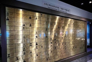 Gold wall with plaques displaying names and portraits, set in a modern room. Korean text at the top highlights a commemorative tone.