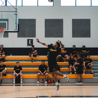 A person in an "ARMY" shirt jumps to hit a volleyball in a gym. Others in similar shirts watch from yellow bleachers. Bright, energetic scene.