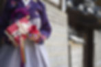 Person in a purple floral hanbok holds a red gift box and colorful pouch. Background shows traditional architecture.