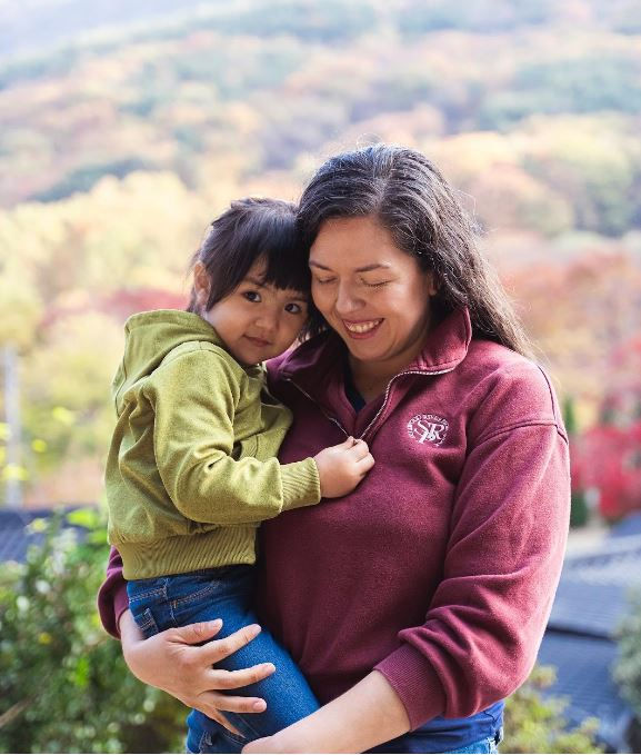 Woman in burgundy holds a child in green. Both smiling, in a vibrant autumn setting with colorful trees. Warm, joyful mood.