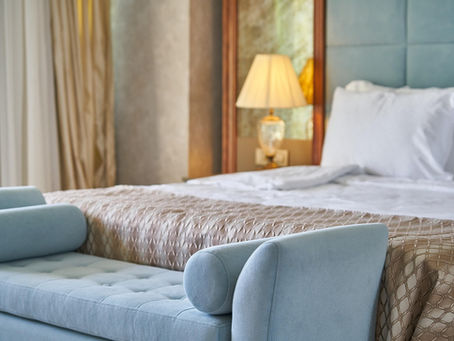 Luxurious bedroom with a blue upholstered bench, beige quilted bedspread, white pillows, and a lit lamp on a bedside table, creating a cozy mood.