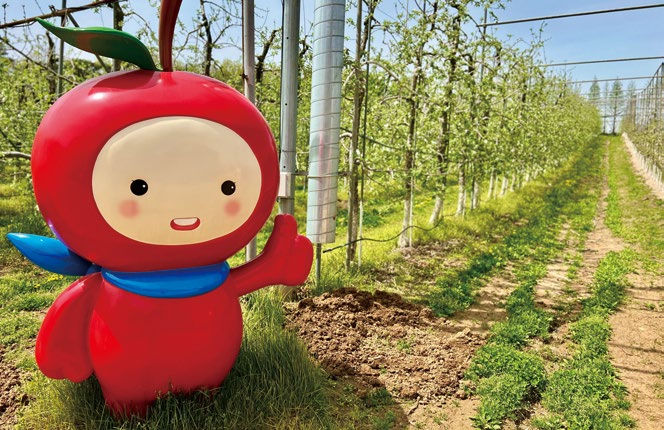 Cheerful red apple character with a green leaf and blue scarf waves in a lush orchard under a clear sky. Rows of trees in the background.
