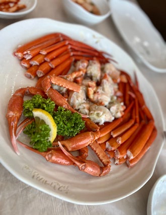 Plate of red steamed crab legs with lemon and parsley garnish on a white table. Empty plates in the background. Elegant dining setting.