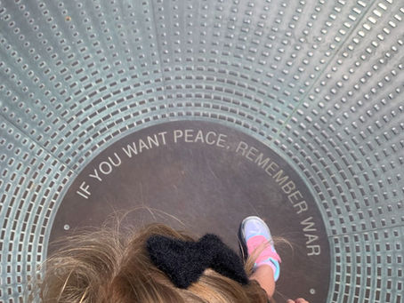Child in pink stands on a circular metal grate with text "IF YOU WANT PEACE, REMEMBER WAR." Sunlight highlights the scene.