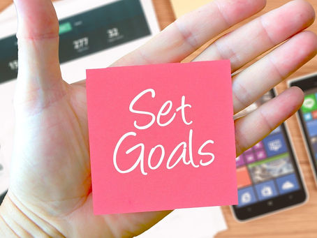 Hand holding pink sticky note with "Set Goals" text. Background shows papers with charts and two smartphones on a wooden table.