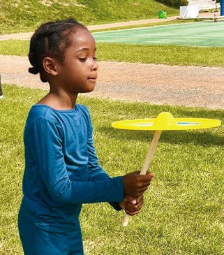 Child in blue outfit spinning a yellow plate on a stick outdoors. Green grass and a track in the background. Focused expression.