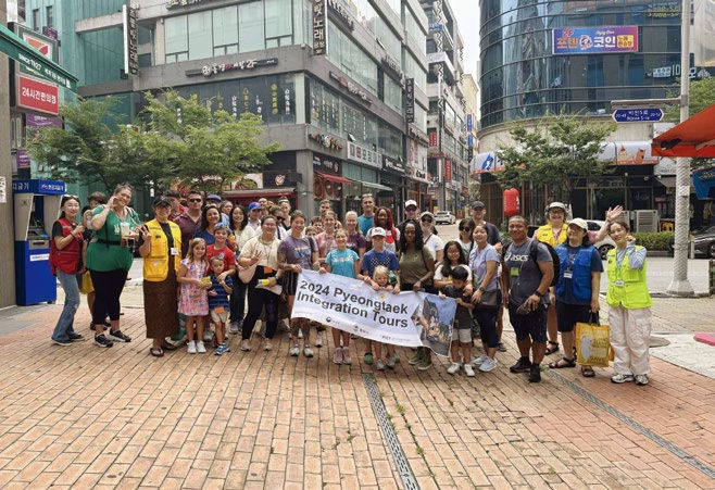 Group of people posing with a "2024 Pyeongtaek Integration Tours" banner in a bustling city street, surrounded by tall buildings and trees.