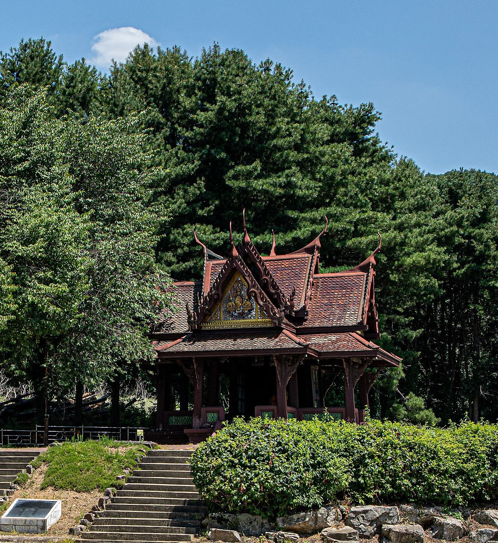 Ornate, traditional pavilion with a decorated roof set amidst lush green trees and shrubbery under a clear blue sky. Stone steps lead up to it.