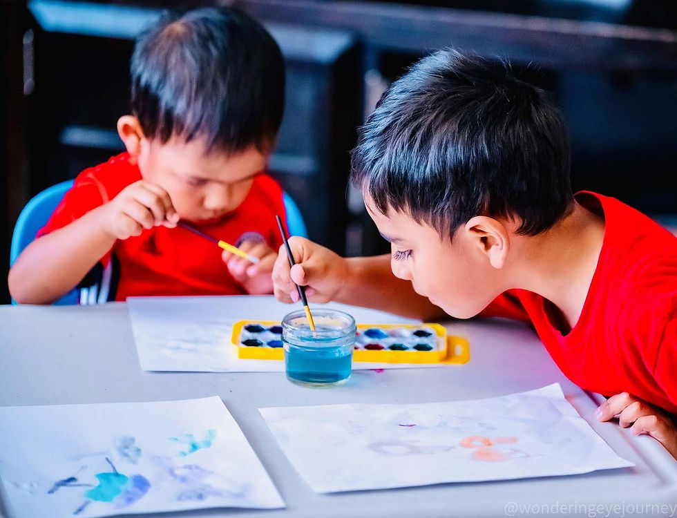 Two boys in red shirts paint with watercolor at a table, focusing intently. Paintings and a palette are visible, creating a playful scene.