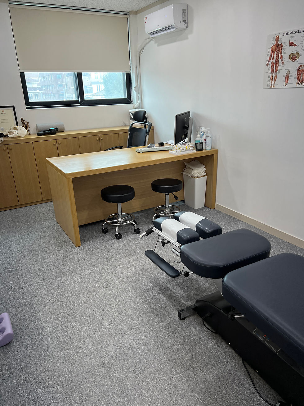 Chiropractic office with a treatment table in the foreground, a wooden desk with stools, and anatomical poster on the wall. Airy, clinical feel.